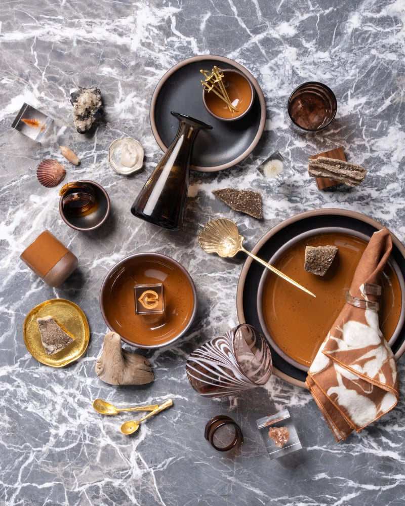 A flat lay of brown ceramic dishes and glassware set on a marble surface, accompanied by decorative elements like seashells, stones, a brown napkin, and gold utensils, creating an earthy and elegant display.