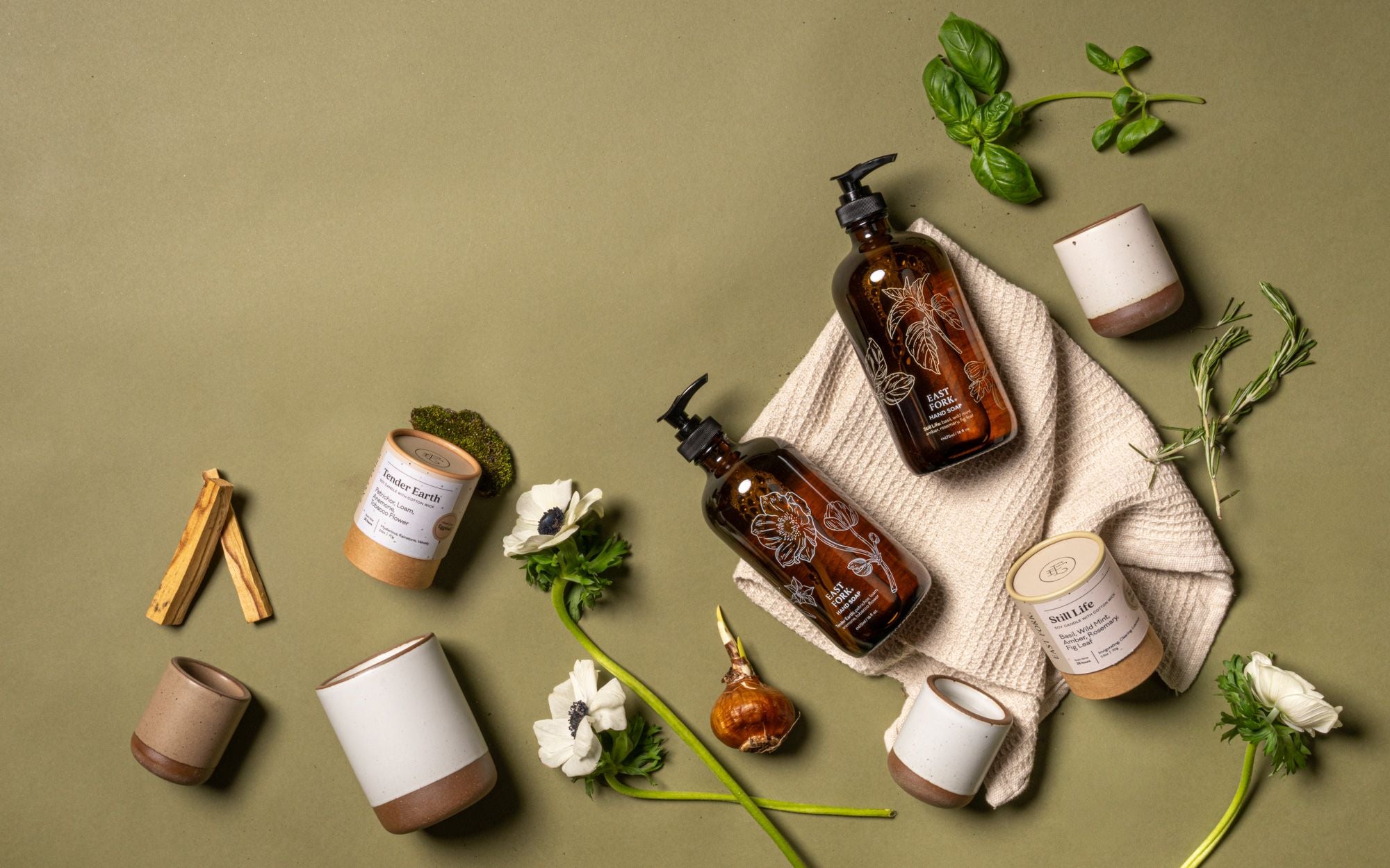 Flat lay of amber soap bottles on a folded towel, ceramic cups, candles, flowers, herbs, and wood sticks on a green background.