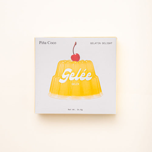A box of Gelée Piña Coco Gelatin Delight, featuring a yellow gelatin mold topped with a cherry on a minimalist white package.