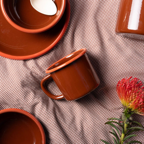 A red-brown enamel mug with an orange rim and handle, laying on fabric with enamel dinnerware in the same colorway and a red flower.