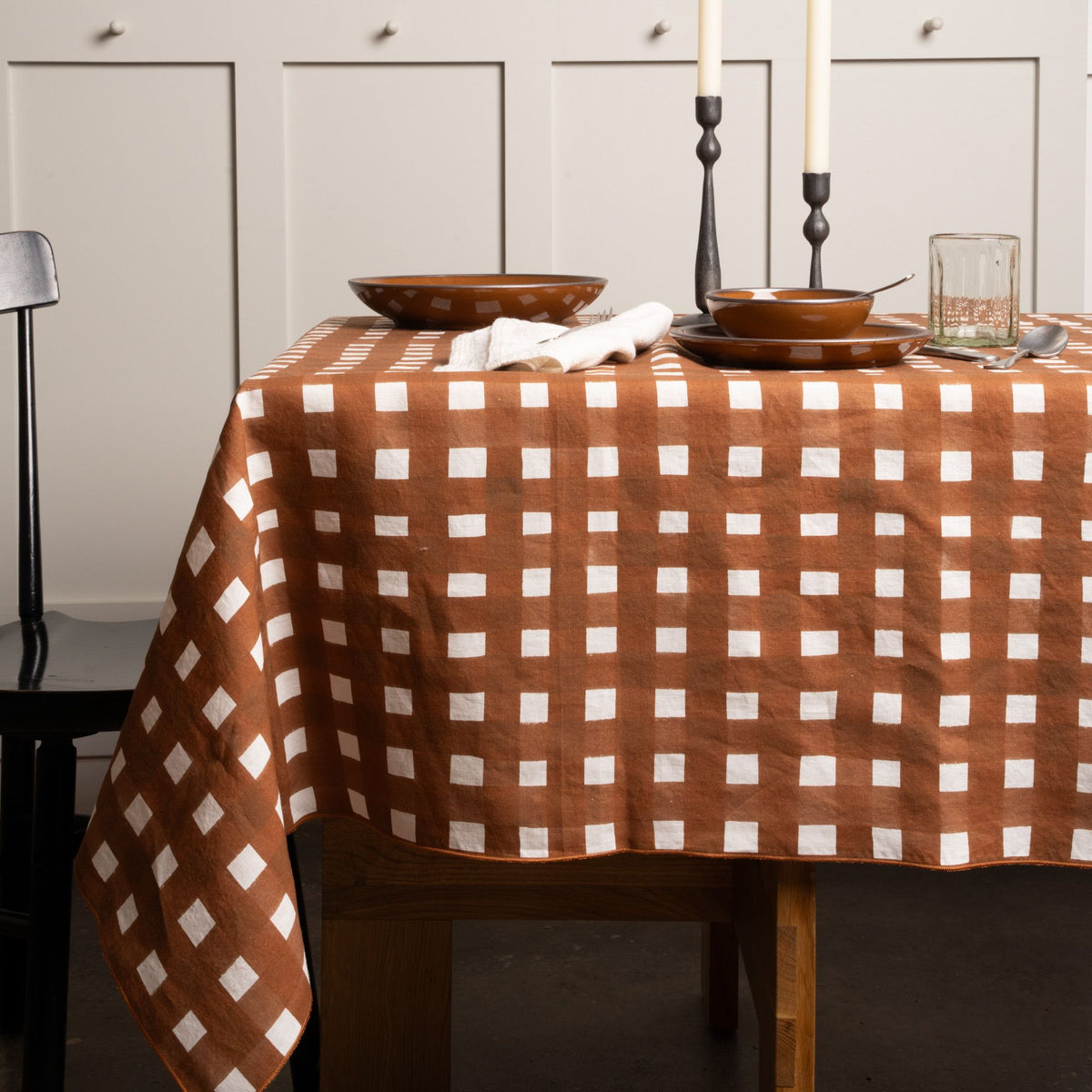 Brown and white crosshatch tablecloth on a dining table, set with bowls, glassware, candles, and a black chair.
