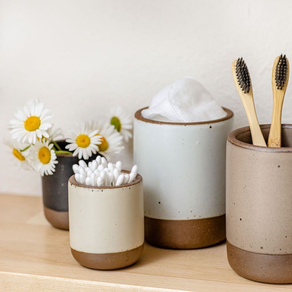 On a birch shelf, there are four candle vessels in various neutral colors, each holding something different: q-tips, toothbrushes, flowers.