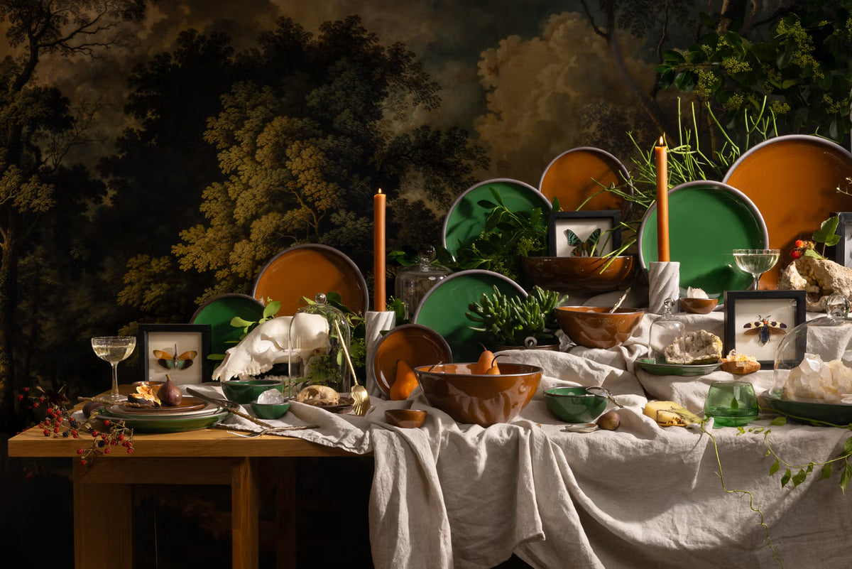 Table with glossy green and brown ceramic plates, candles, crystals, plants, and framed insects against a forest mural backdrop.