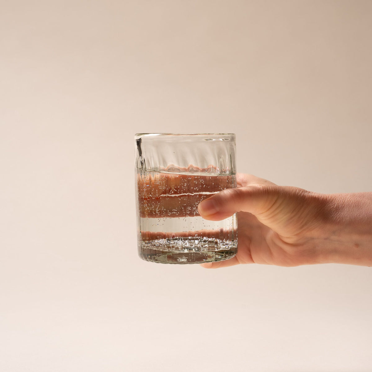 A hand holds out a taller clear glass tumbler with vertical ridges, filled with water, set against a neutral background.