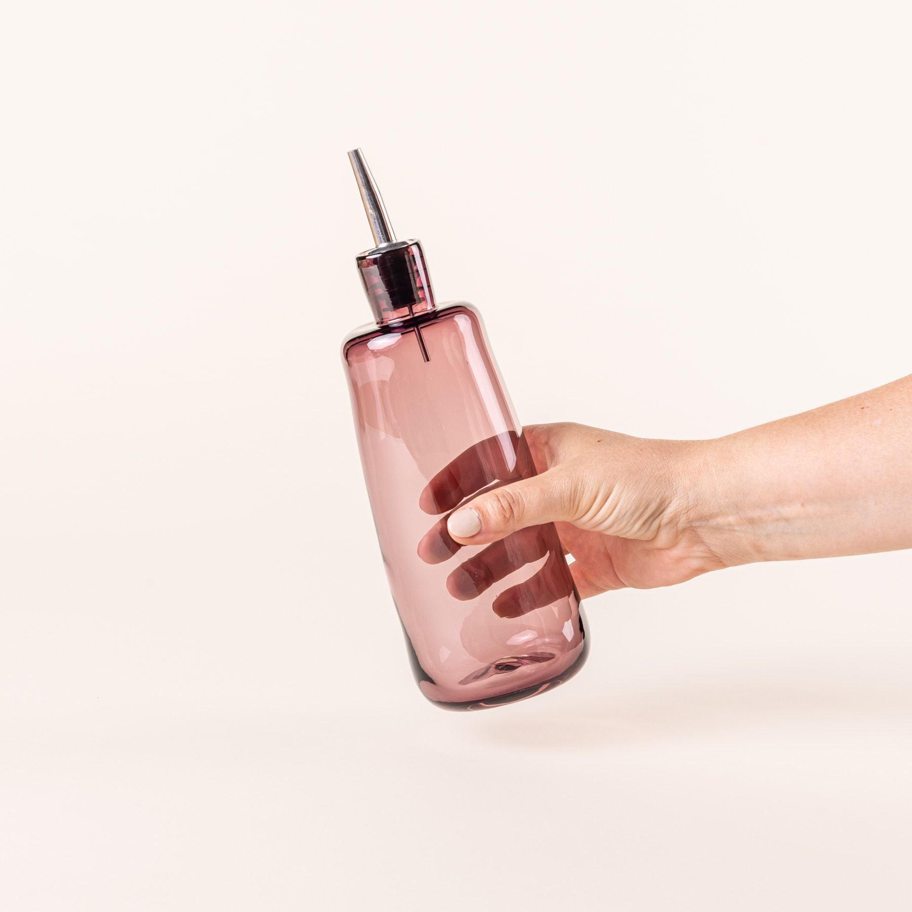 A hand holds a tall glass cylindrical cruet in a light plum color
