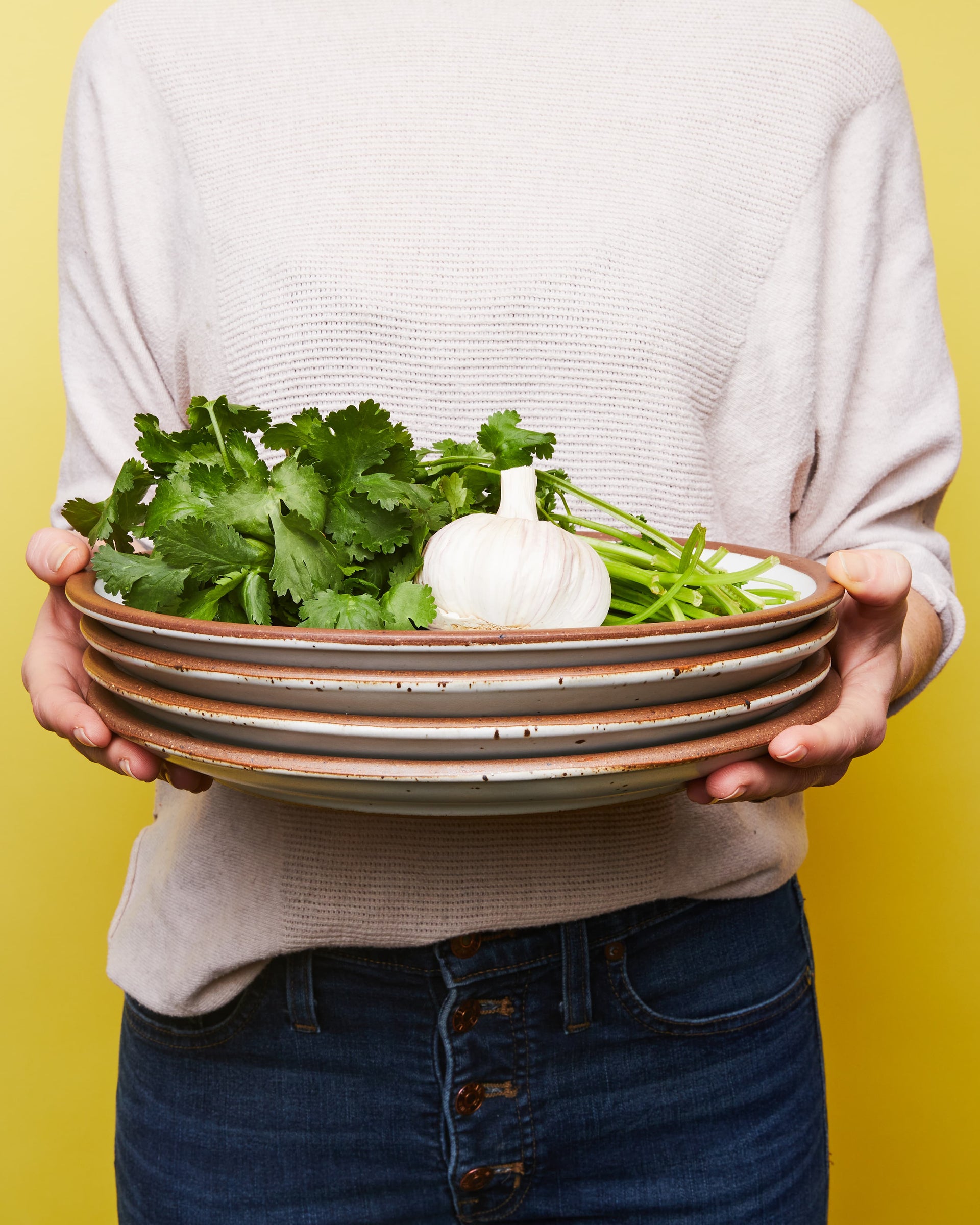 A person holds a stack of white ceramic dinner plates, topped with herbs and a whole garlic.