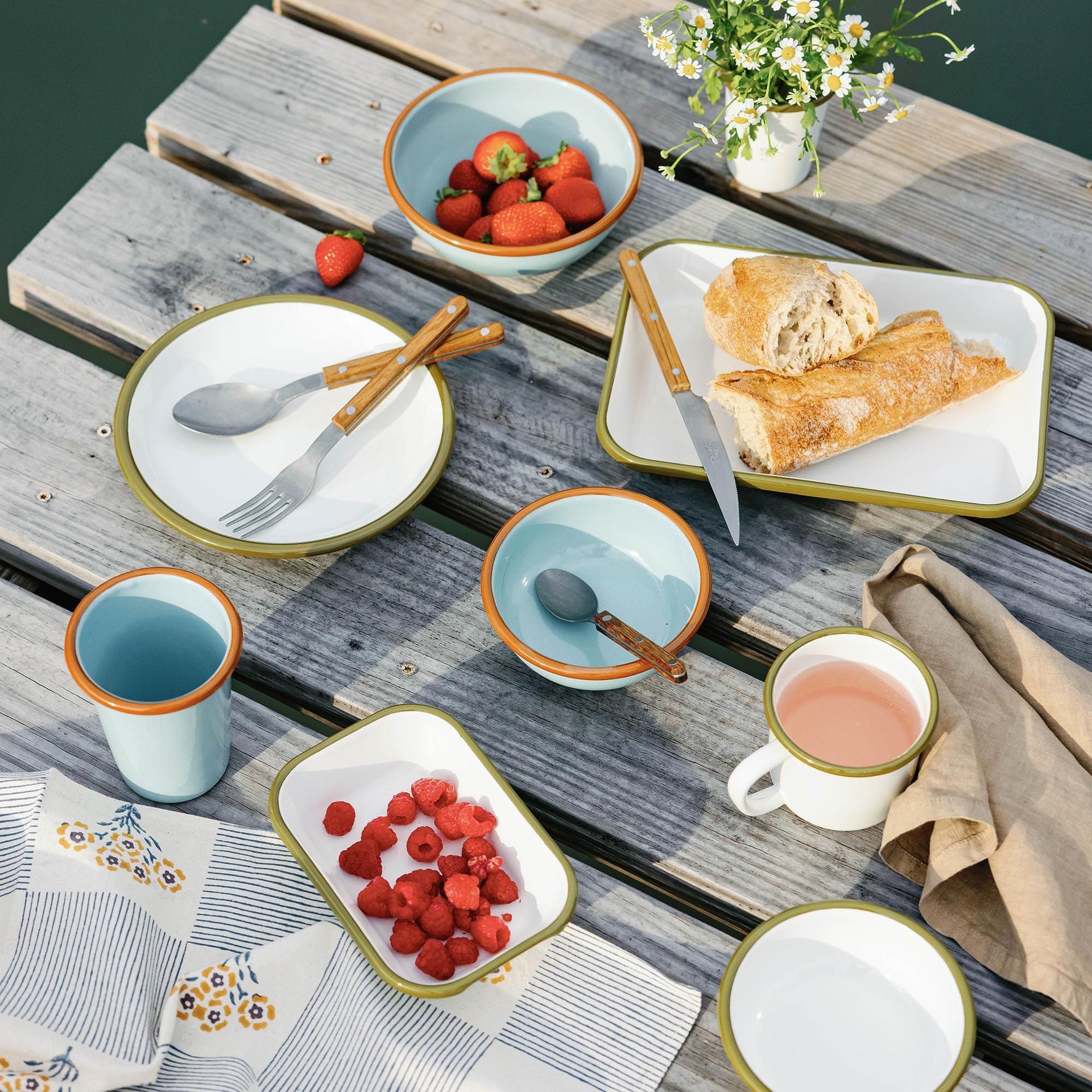 Outdoor picnic spread with enamel dishes, berries, bread, and pastel cups arranged on a weathered wooden dock.