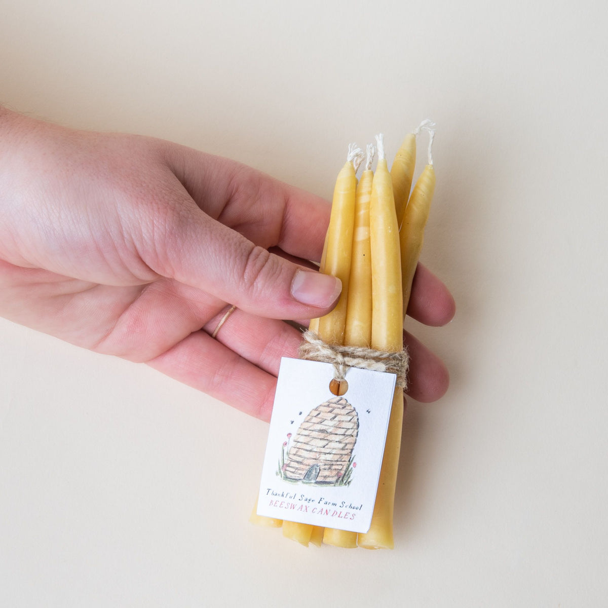 A hand holding a little bundle of short taper beeswax candles with string tied around them with a white packaging tag