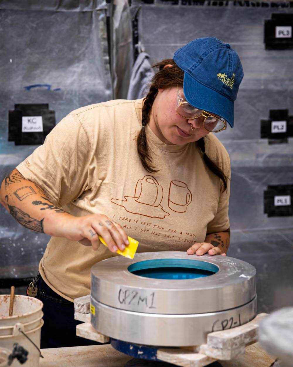 A person in a beige T-shirt with mug illustrations and a blue cap works in a ceramics studio, using a tool on a metal mold labeled "CP2 M1". The background includes a shelf with organized items and a bucket with pottery tools.