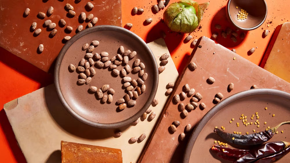An overhead view of plates and tiles in a warm brown color with ingredients like dried pinto beans, peppers, seeds, and a tomatillo