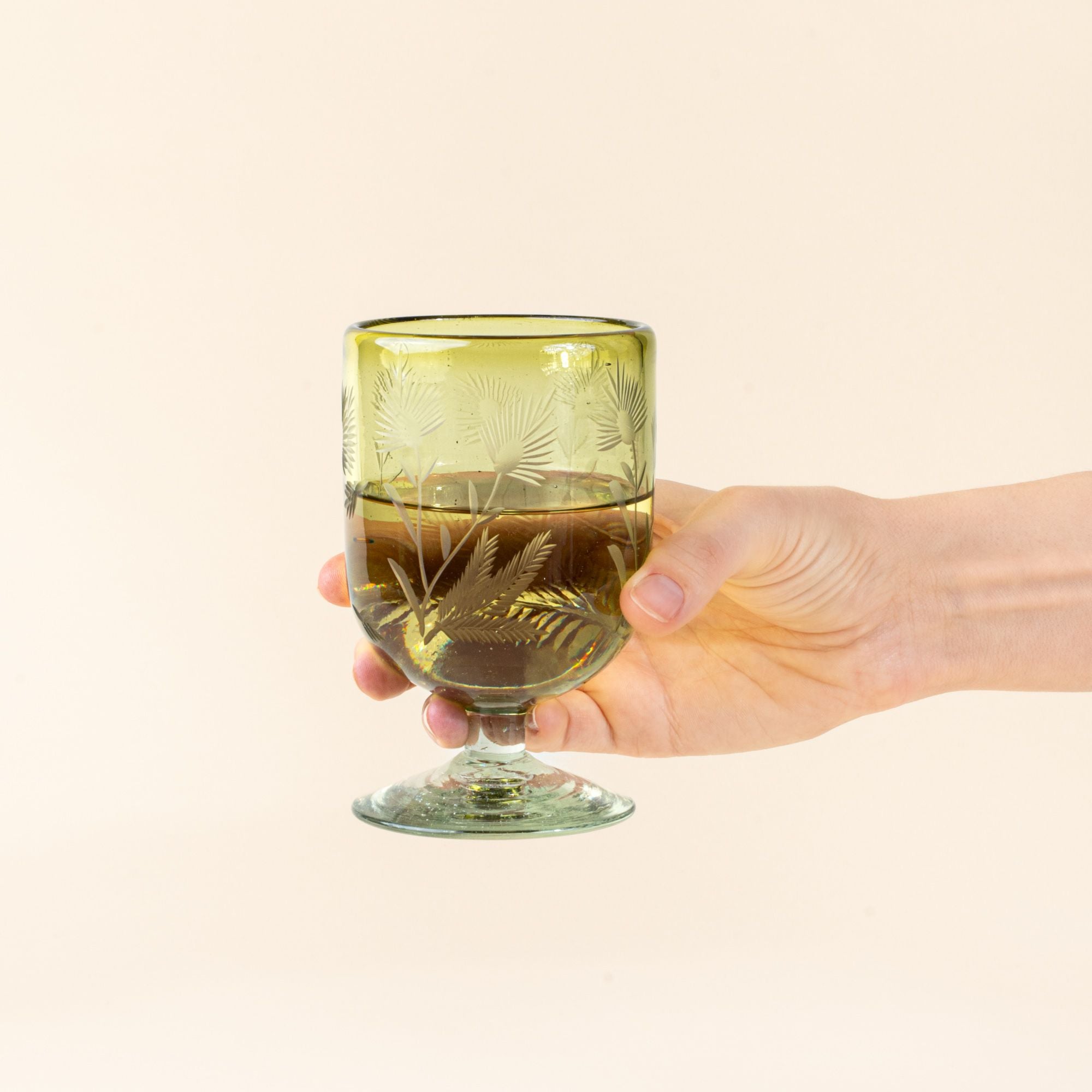 A hand holds out an olive short wine glass made of recycled glass with etched thistle designs on it, filled with white wine.