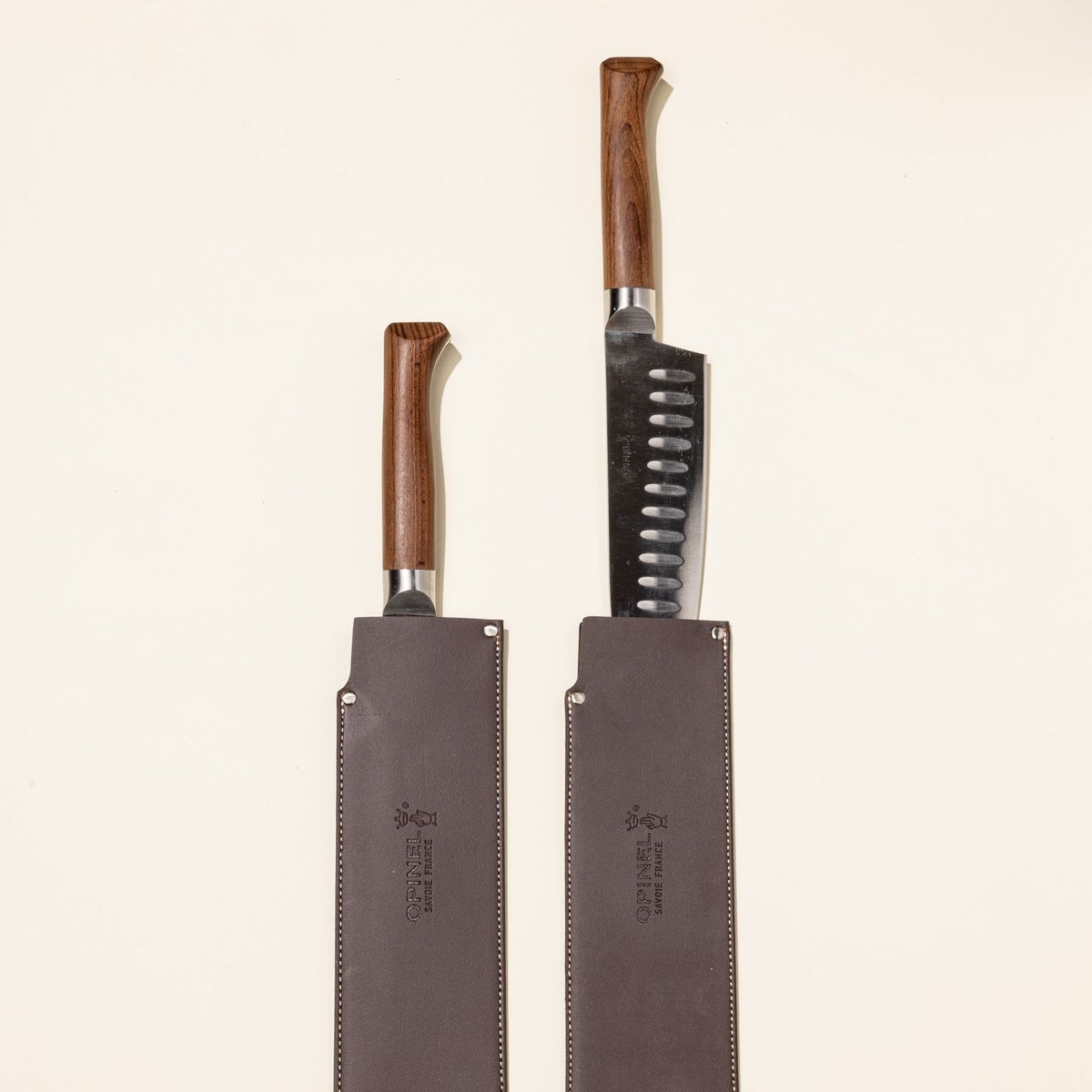 A santoku knife with a sharp steel blade and a beveled dark wood handle sits with a leather sleeve on the blade. Next to it is another knife sitting fully inside a leather sleeve.