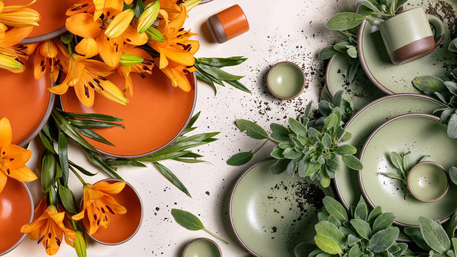 Orange and green ceramic plates and mugs are artfully arranged with vibrant orange lilies and soft green lamb's ear leaves. Soil is scattered across the light surface, adding a natural, earthy element.