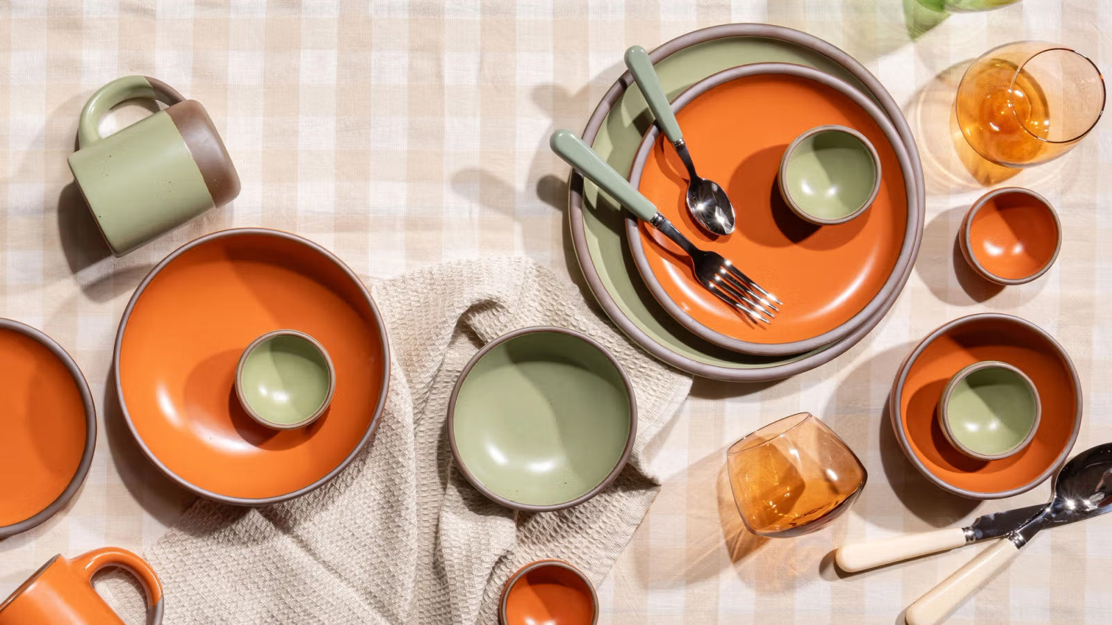 A vibrant table setting featuring East Fork pottery in daylily and green hues, including plates, bowls, mugs, and glassware, arranged on a checkered tablecloth with utensils and textured napkins.