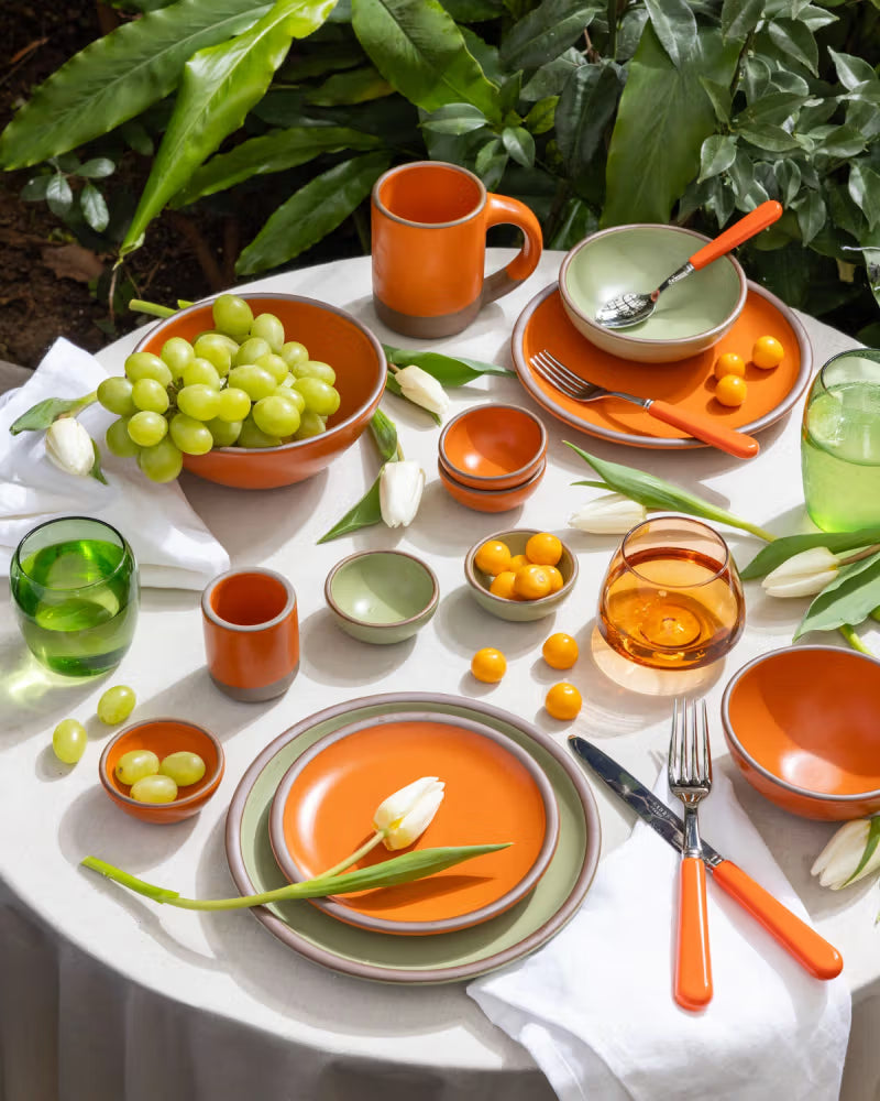 A vibrant outdoor table setting featuring East Fork pottery in Daylily and Lamb's Ear glazes, including mugs, bowls, plates, and small dishes. The table is adorned with green grapes, small orange fruits, white tulips, and glassware, surrounded by lush green plants.