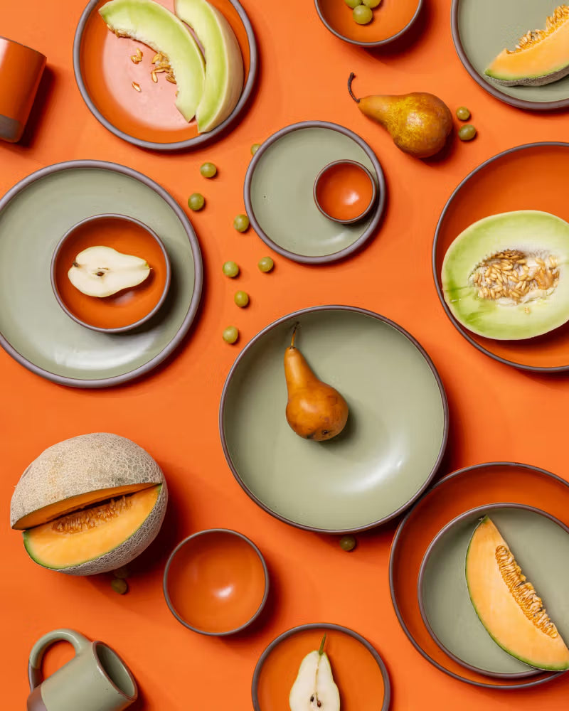 Green and orange ceramic dishes, bowls, and mugs are arranged on an orange surface alongside sliced melons, pears, and scattered green peas.