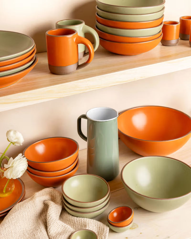 Ceramic dinnerware in orange and green glazes, including plates, mugs, and bowls, are displayed on wooden shelves. A textured cloth and a small vase with white flowers add to the arrangement.