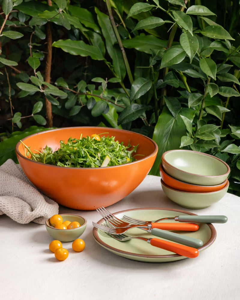 A vibrant tablescape featuring a large orange bowl filled with salad, a stack of green bowls, and matching plates with green and orange handles, set against a backdrop of lush green foliage.