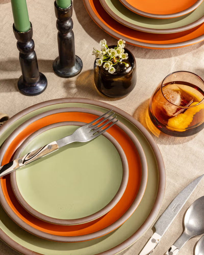 A set of plates in orange and green glazes is arranged on a linen tablecloth alongside a fork and knife. Two dark candlesticks with green candles and a small vase with white flowers accompany a glass of iced tea with lemon.