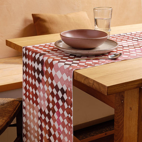A wooden dining table styled with a pink patterned table runner, pink and warm pale brown ceramic plates and bowls, silver utensils.