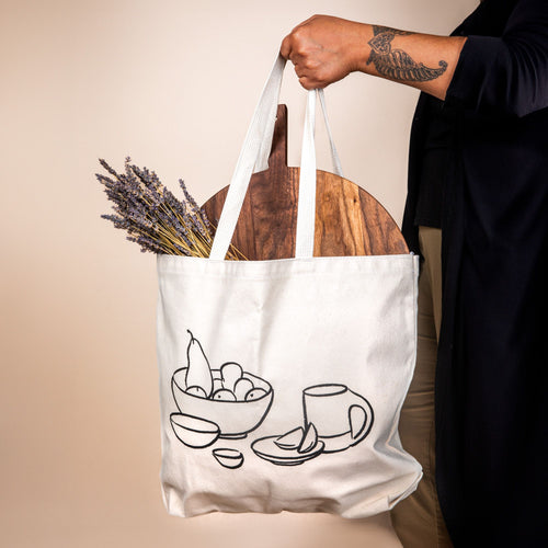 A cream canvas tote has a charcoal illustration on the front of a bowl with fruit, little bowls, plate, and a mug. A hand holds the tote and a large serving board and bundle of lavender is hanging out of the top.