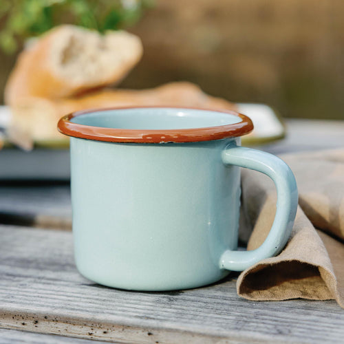 Light blue enamel mug with a brown rim on a weathered wooden table, napkin and sliced bread in background.