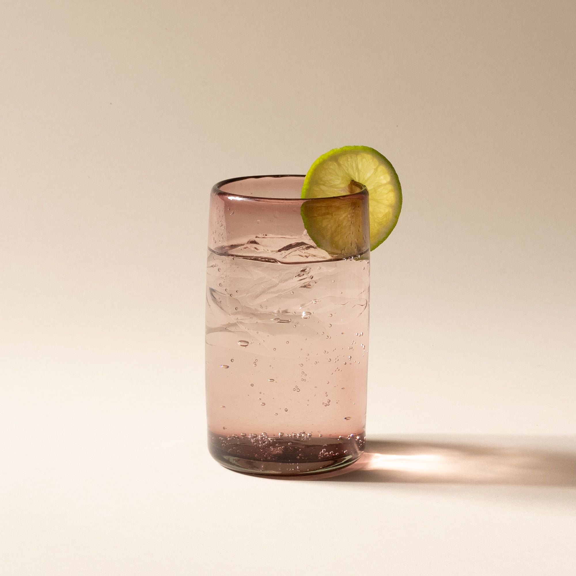 A cocktail with a lime, raspberry, and mint garnish in a light plum tall transparent drinking glass
