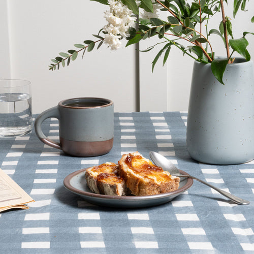On a blue crosshatch tablecloth sits a cake plate in a blue-grey color with toast and spoon, nearby is a small mug and vase.