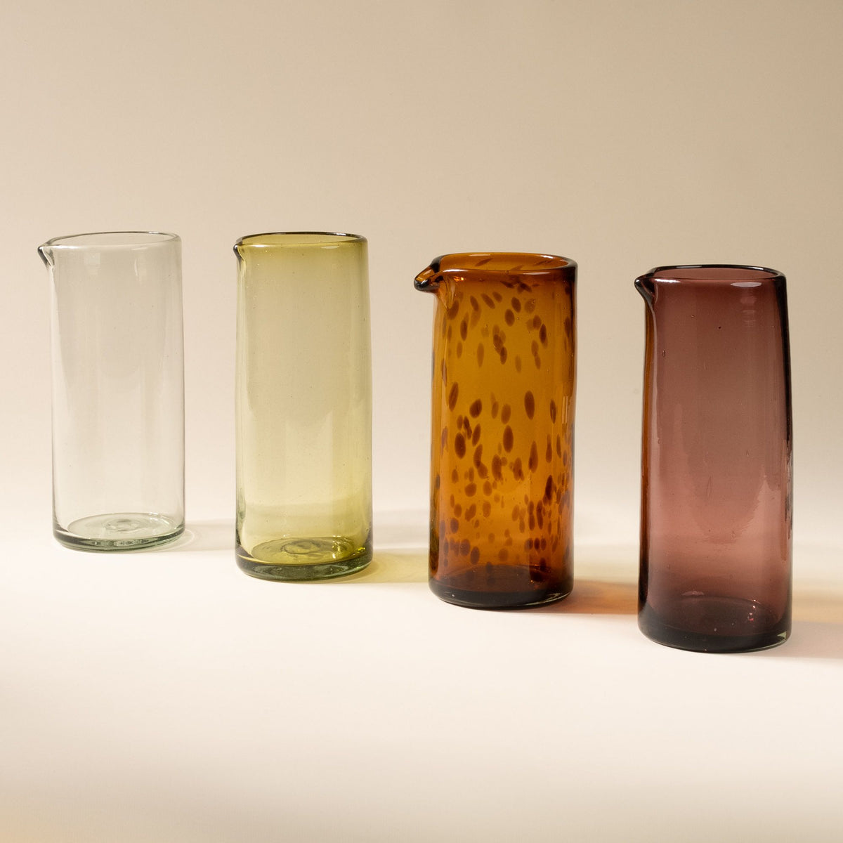 A row of tall transparent glass pitchers in different colors including: tortoiseshell, light plum, olive green, and clear