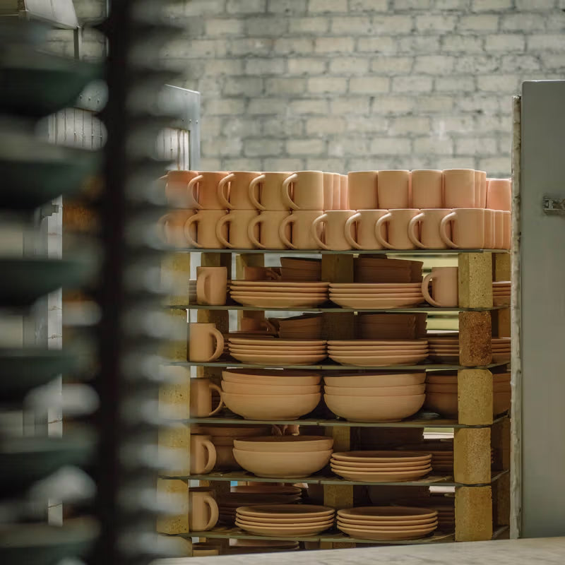Stacks of unglazed ceramic mugs, bowls, and plates in a kiln, with a brick wall in the background, preparing for firing.