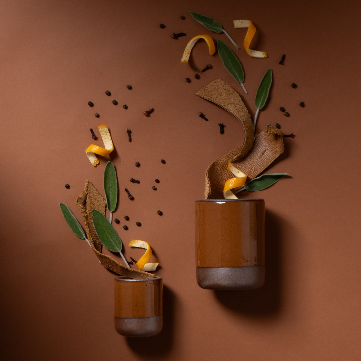 Two brown ceramic tumblers with orange peel, sage leaves, and spices arranged artistically on a brown background.