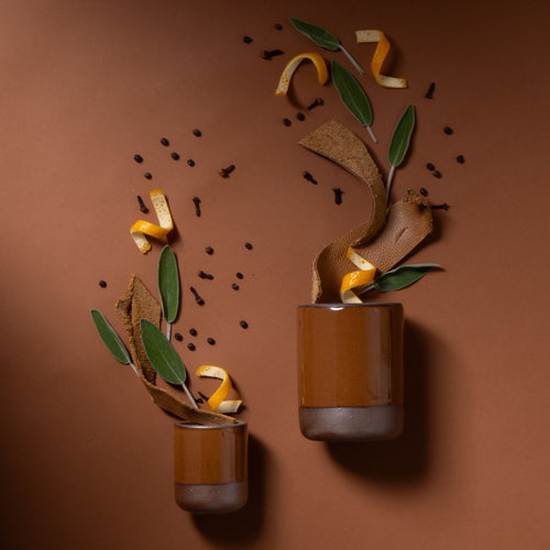 Two brown ceramic tumblers with orange peel, sage leaves, and spices arranged artistically on a brown background.