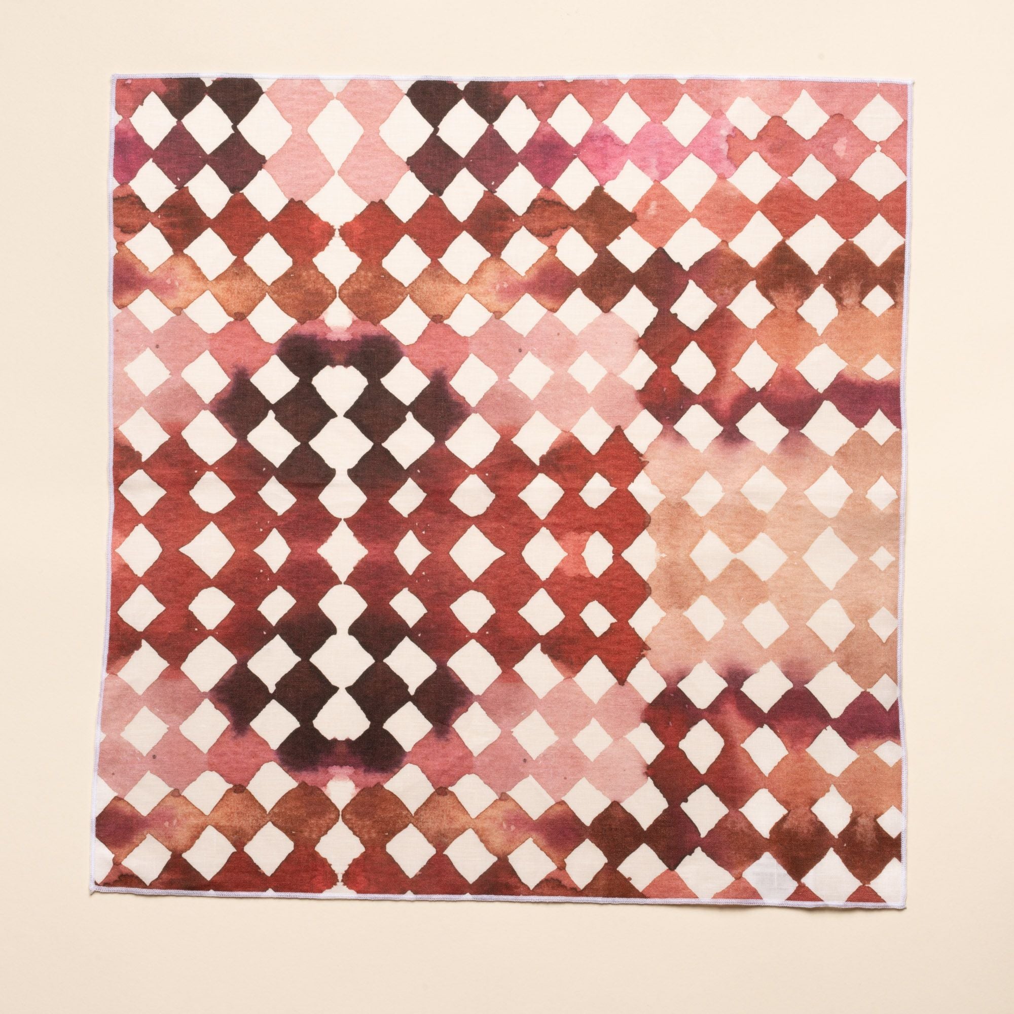 A flat square watercolor diamond-patterned cloth napkin in warm burgundy and pink tones
