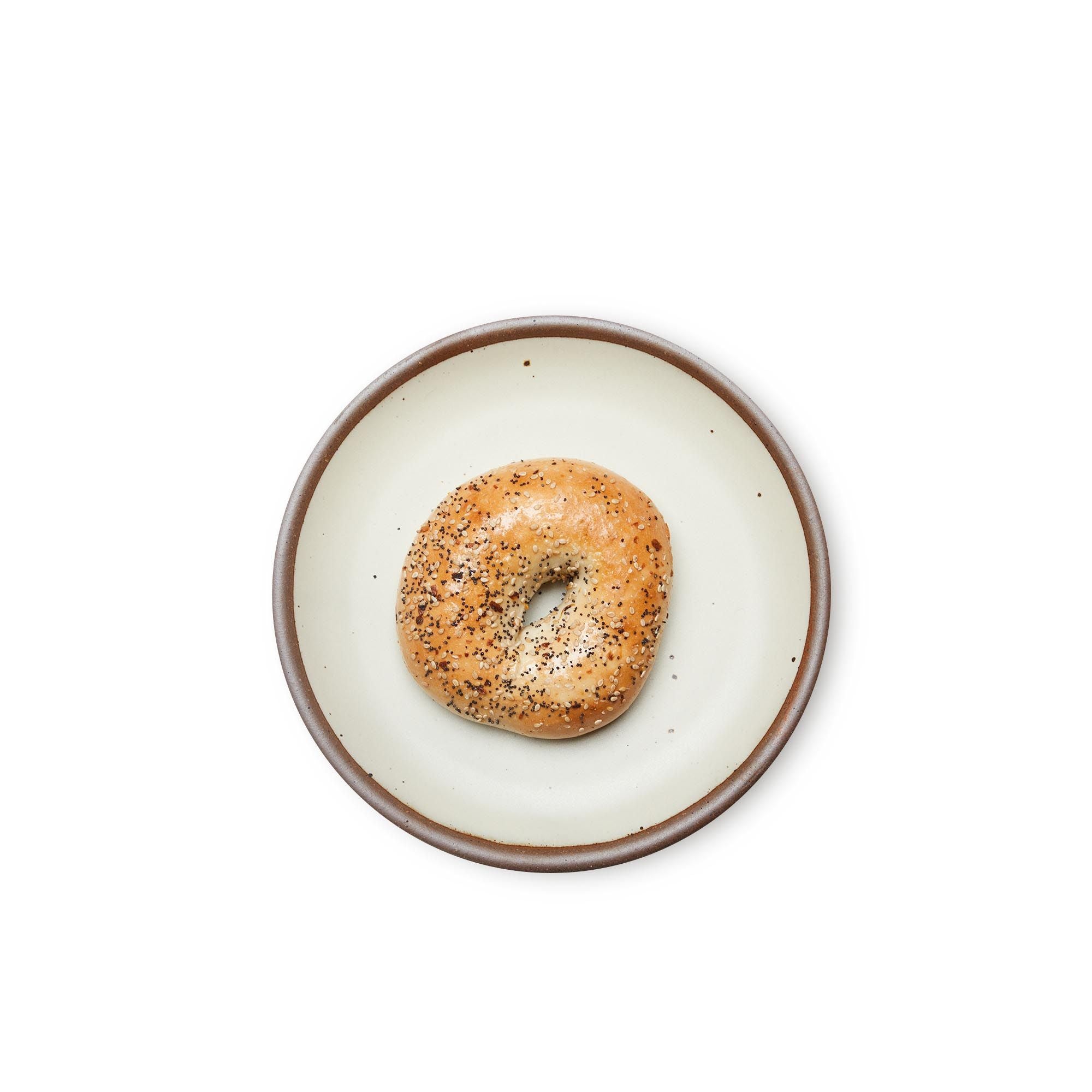 A bagel on a medium sized ceramic plate in a warm, tan-toned, off-whitecolor featuring iron speckles and an unglazed rim.