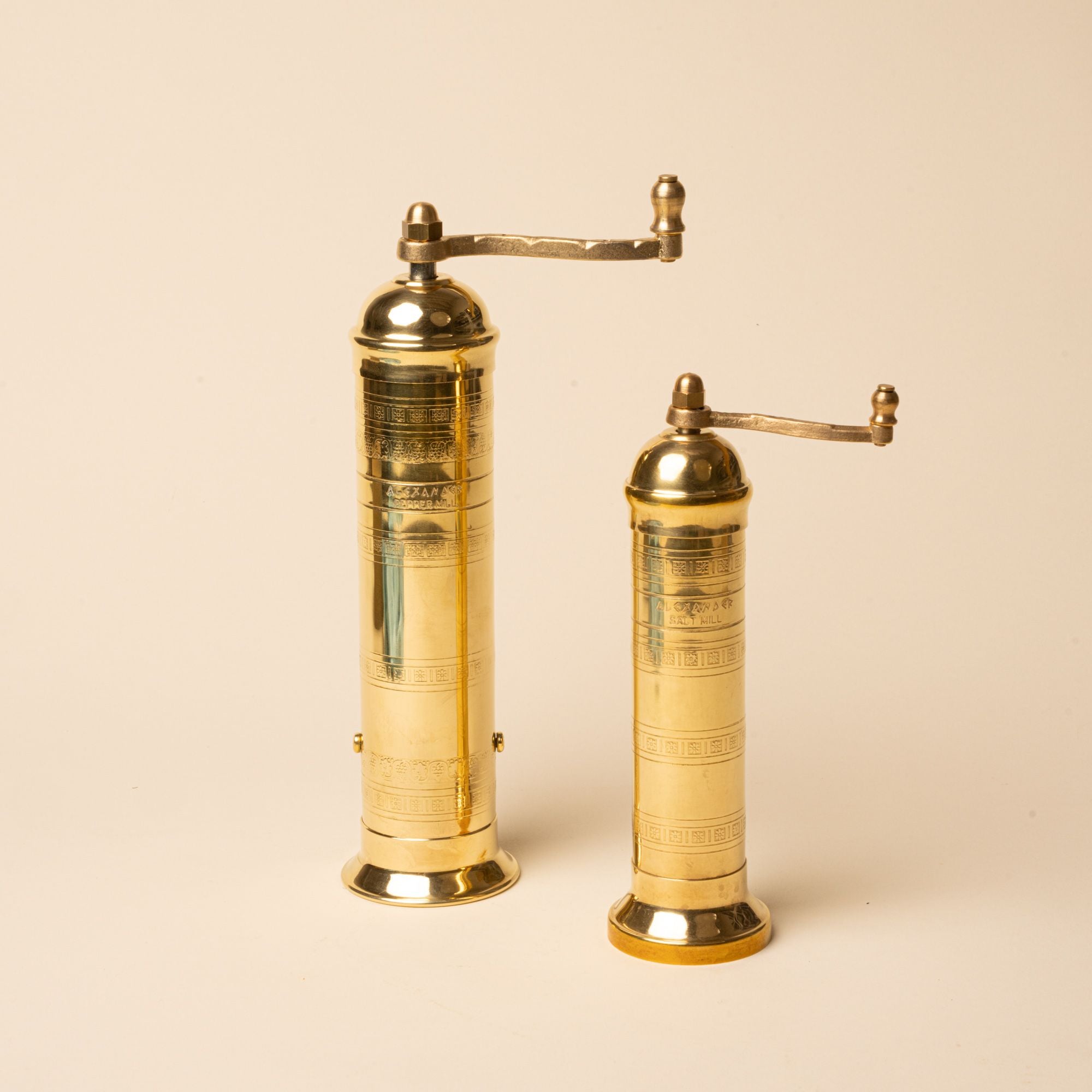 Two brass hand-crank salt and pepper mills of different sizes stand side by side on a neutral background.