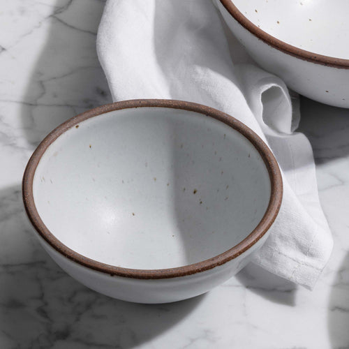 A small dessert sized rounded ceramic bowl in a cool white color featuring iron speckles with a napkin of the same color.