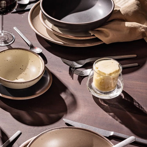 A table filled with place settings of ceramic dinnerware and silverware, and a clear glass butter keeper filled with light yellow butter