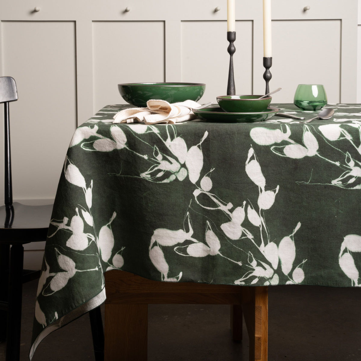 Dark green tablecloth with white botanical print, set with green dishes, candles, and glassware on a wooden table.