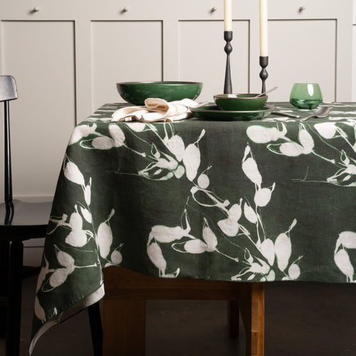 Dark green tablecloth with white botanical print, set with green dishes, candles, and glassware on a wooden table.