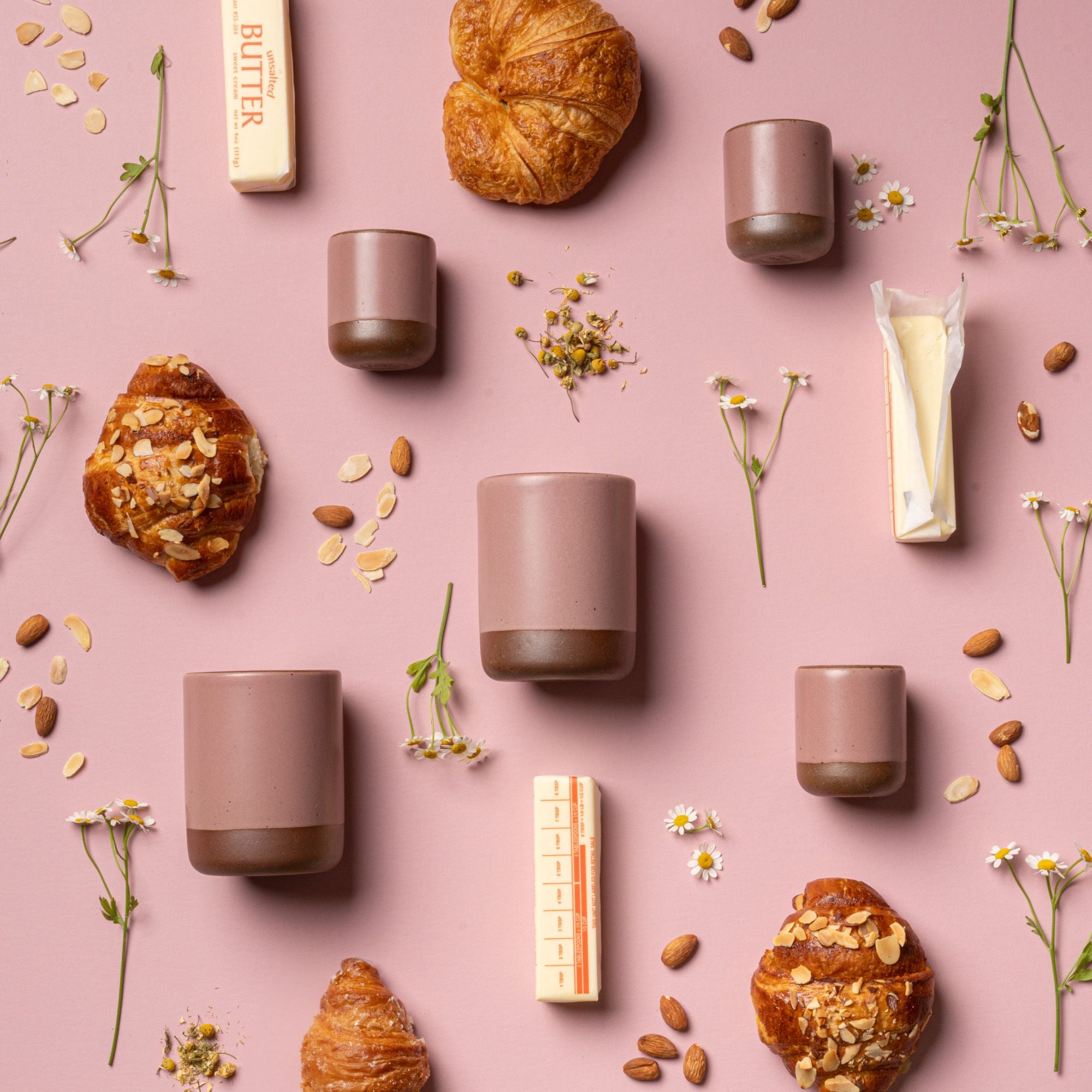 An artful layout of The Candle in Almond Flower - small and large ceramic candles in a cylindrical vessel in a soft mauve pink color, surrounded by almonds butter, and tiny flowers.