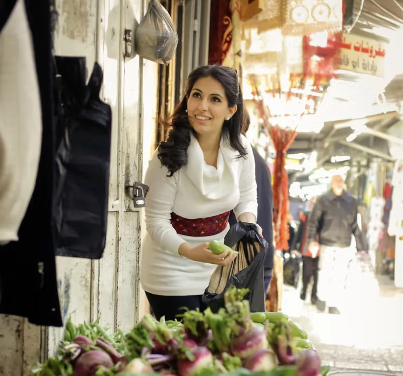 A woman in a white top with a red belt is at a market stall, holding a black bag and a vegetable. She is surrounded by fresh produce, and other shoppers are visible in the background.