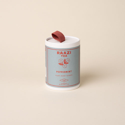 A white cylindrical tin of RAAZI Tea labeled ‘Peppermint’ with a muted blue label and red text, featuring a red pull tab on the lid, photographed on a pale beige background.