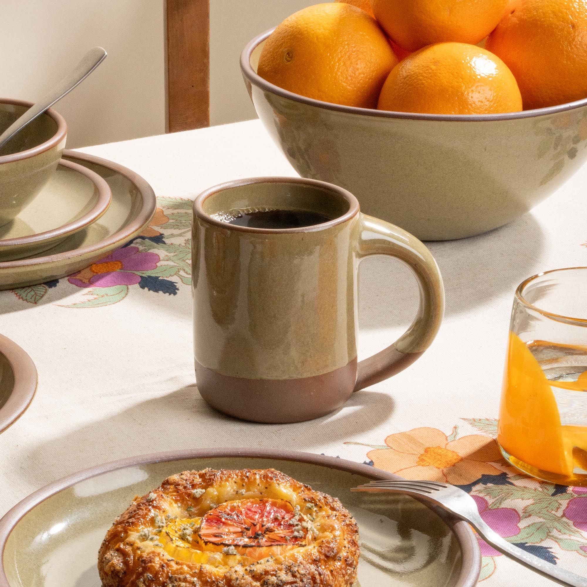 Coffee in a stoneware grey-green mug beside citrus, pastries, and stacked plates on a floral tablecloth.