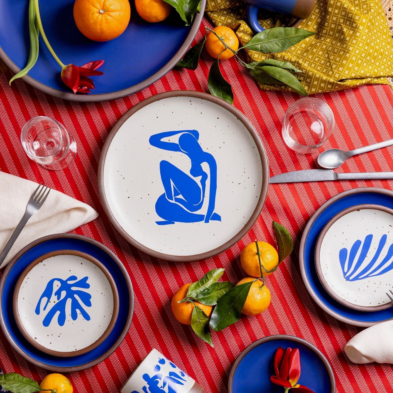 On a colorful table are plates in a bright cobalt blue, with decorated plates featuring artwork by Henri Matisse, along with silverware, glasses, and oranges..