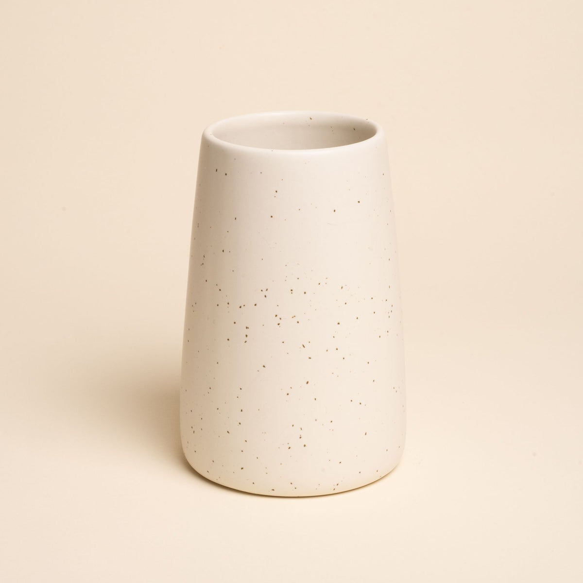 A large minimalist cream tumbler vase with subtle iron speckles stands centered on a soft beige background