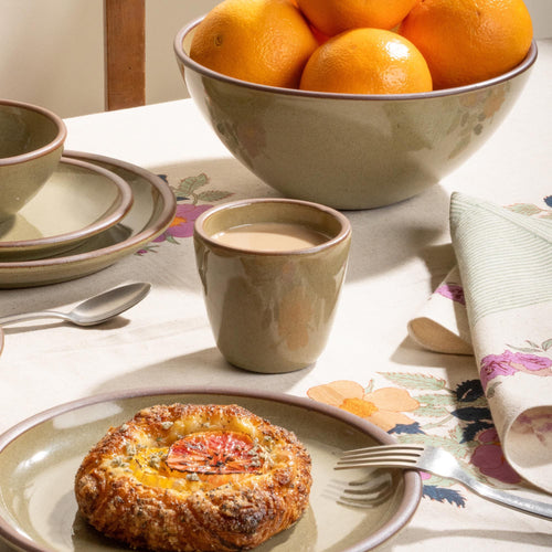 Grey-green stoneware tapered cup with cream coffee, oranges, and pastry on a floral table setting.