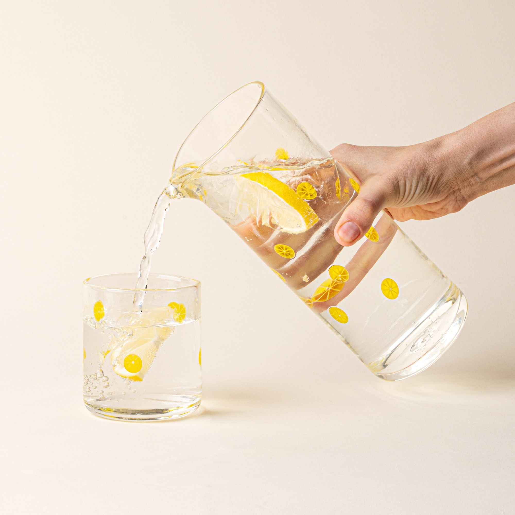 A hand holding a clear glass pitcher with yellow lemon slice motif and pour spout, pouring water into a matching cup.