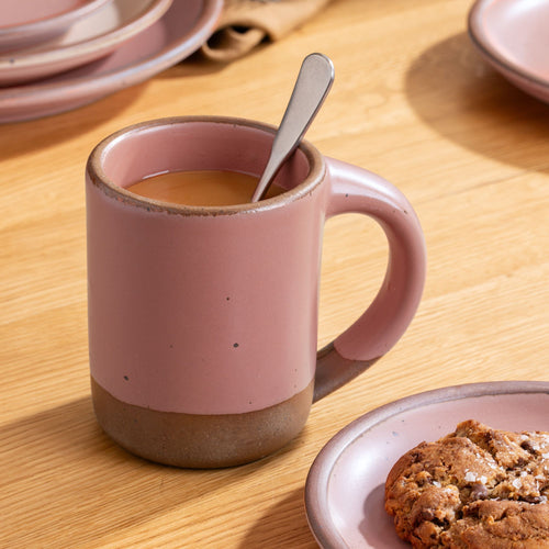 Dusty mauve pink ceramic mug with coffee and a spoon, set on a wooden table beside a cookie on a matching plate.