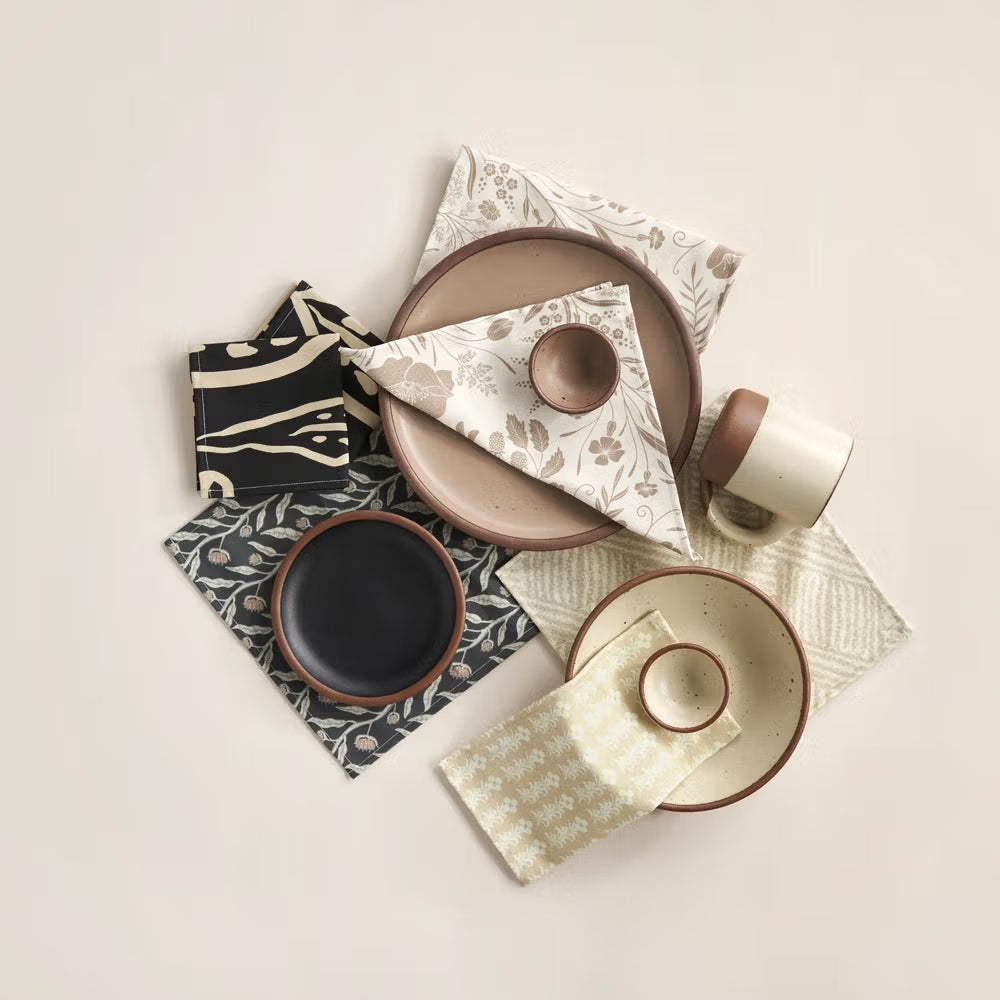 An assortment of ceramic dinnerware and textiles arranged artfully. The collection includes plates, bowls, and a mug in neutral and clay tones, alongside patterned napkins with floral and geometric designs on a light background.