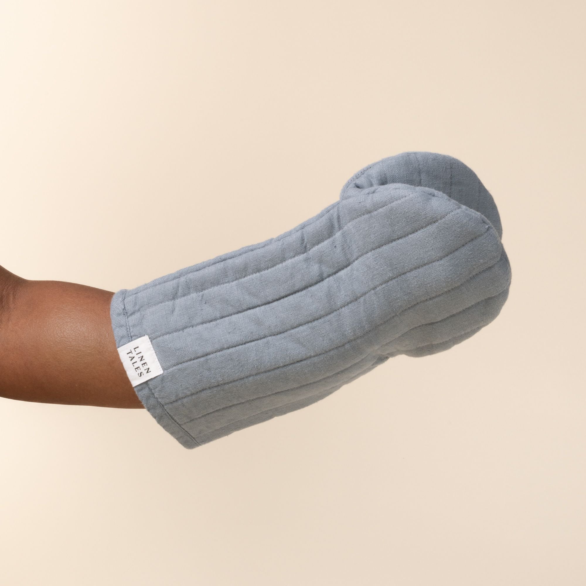Hand inside a soft blue linen oven mitt with stitched channels and a small ‘Linen Tales’ label, against a light background.